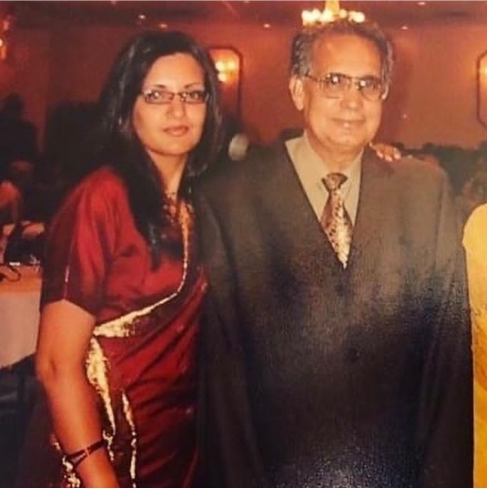 A woman wearing a red saree stands beside an older man in formal attire, both smiling at the camera. The background features an event setting with soft lighting.